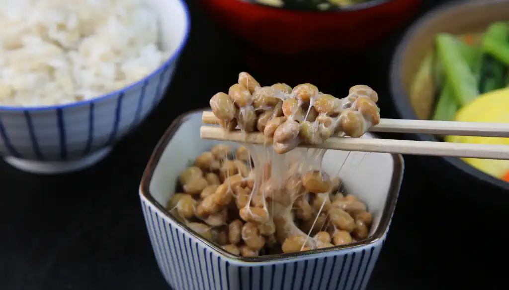 Nattokinase ist ein traditionelles japanisches Gericht aus fermentierten Sojabohnen, die klebrig und fadenbildend sind. Der Geschmack und Geruch sind stark und unverwechselbar. Oft wird Natto mit Reis serviert und kann mit gehackten Frühlingszwiebeln, Senf oder Sojasauce verfeinert werden. Beim Essen zieht sich die klebrige Konsistenz in Fäden, die mit Stäbchen aufgerollt werden. Die Reaktion der Essenden variiert von Genuss bis Skepsis, je nach Vertrautheit mit dem Geschmack. Die Szene spielt oft in einer traditionellen japanischen Umgebung, z.B. an einem niedrigen Tisch auf Tatami-Matten.